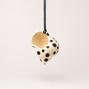 May include: A ceramic shell-shaped pendant in a cream color with hand-painted black polka dots. The pendant is suspended from a thin black cord, featuring a hollow interior and a smooth, glossy finish that highlights the rounded, spiral form.