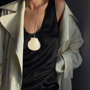 Puede incluir: Un collar con colgante de concha de color crema cuelga sobre una blusa negra. El colgante es una concha de vieira grande y detallada. Una gabardina beige claro está drapeada sobre los hombros.
