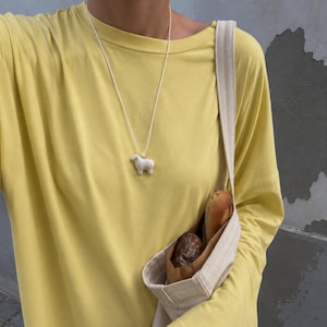 May include: A white sheep pendant necklace hangs over a yellow long-sleeve shirt. A beige canvas bag holds two loaves of bread. The background is a gray wall.
