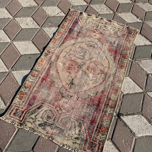 May include: A rectangular, distressed rug with a faded red and beige central design. The rug features a central medallion and ornate border details in red, green, and beige. The rug is placed on a geometric patterned surface.