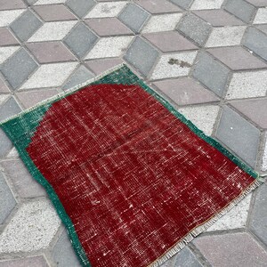 Puede incluir: Una alfombra rectangular y desgastada con un centro rojo y un borde verde. La alfombra tiene un aspecto descolorido y vintage con desgaste visible. La alfombra está colocada sobre una superficie de ladrillos grises y marrones estampados.
