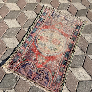 Puede incluir: Una pequeña alfombra rectangular con un aspecto vintage y desgastado. La alfombra presenta un diseño central en tonos rojos, azules y verdes, con un aspecto descolorido y envejecido. Los bordes están bordeados con un patrón detallado y flecos.