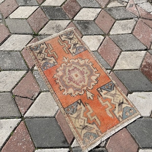 May include: A vintage Turkish rug with a geometric pattern in shades of orange, blue, and beige. The rug has a fringe border and is lying on a gray and brown tiled floor.