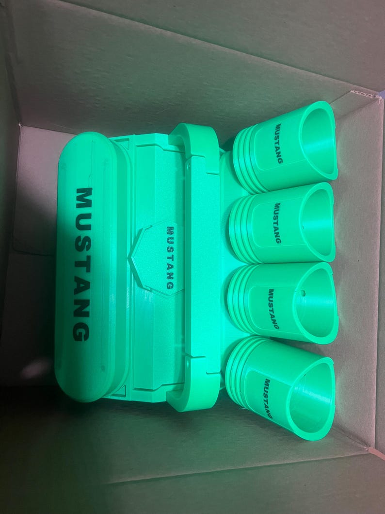 Pode incluir: Um conjunto de quatro copos de shot de pl&aacute;stico verde com a palavra "Mustang" impressa em cada um. Os copos est&atilde;o dispostos em fila, com a palavra "Mustang" voltada para a frente. Os copos est&atilde;o em uma caixa de papel&atilde;o.