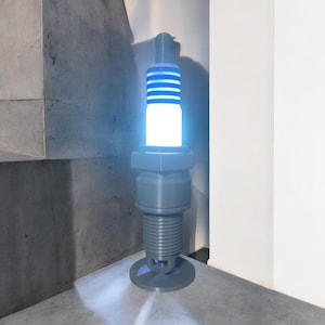 以下が含まれることがあります： 白く光る灰色と青色のスパークプラグの形をしたランプ。ランプは灰色の表面の上に置かれています。