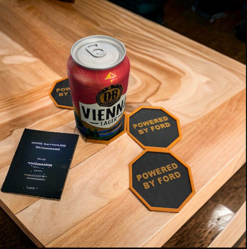 Pode incluir: Uma lata de cerveja Vienna Lager da cervejaria DB, sentada em uma mesa de madeira com dois  apoios de copo pretos e laranja que dizem "Powered by Ford".