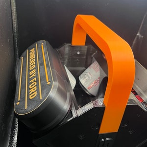 Pode incluir: Uma caixa de armazenamento de pl&aacute;stico impressa em 3D de cor preta e laranja com uma al&ccedil;a. A caixa tem uma etiqueta que diz "Powered by Ford".