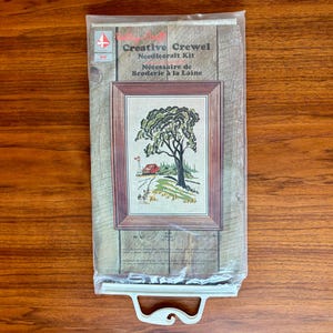 Peut inclure: Un kit de broderie "Creative Crewel" vintage dans un emballage plastique transparent. Le kit comprend une broderie encadrée d'un arbre, d'un train rouge et d'un paysage herbeux. Le paquet contient du texte en anglais et en français.