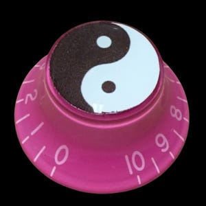 May include: A circular pink control knob featuring a black and white yin-yang symbol on the top surface. The sloped side of the knob displays white numerical markings ranging from zero to ten, with additional tick marks between the numbers.