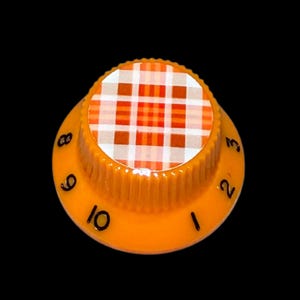 May include: A round orange control knob featuring a ribbed side and a flat top surface with a white, orange, and brown plaid pattern. The outer edge of the knob is marked with black printed numbers ranging from one to ten in a circular sequence.