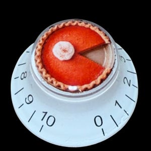 Puede incluir: Un temporizador de cocina redondo con base blanca y números negros del cero al diez. La parte superior del temporizador muestra un gráfico de una tarta de calabaza naranja con un copete de nata blanca y una porción triangular faltante.