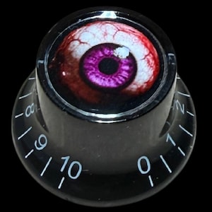 Puede incluir: Un mando de control cilíndrico negro con un gráfico de un ojo humano realista con un iris púrpura vibrante y esclerótica roja inyectada en sangre en la parte superior. El lateral del mando está marcado con números blancos del cero al diez.