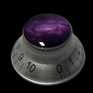 Puede incluir: Perilla de control plateada con un diseño de galaxia. La parte superior es de color púrpura oscuro con patrones blancos y morados claros. La perilla tiene números del 0 al 10 alrededor de la base.