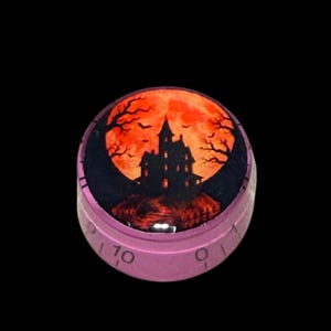 Peut inclure: Un bouton de commande circulaire violet avec un insert graphique représentant la silhouette noire d'une maison hantée sur une grande pleine lune orange vif. Le côté du bouton est marqué d'incréments numériques noirs, dont le chiffre 10.