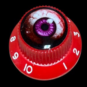 Pode incluir: Um botão de controlo circular vermelho com uma borda estriada e números brancos impressos de um a dez. O centro do botão apresenta um gráfico detalhado de um olho humano realista com uma íris roxa vibrante e veias vermelhas visíveis.