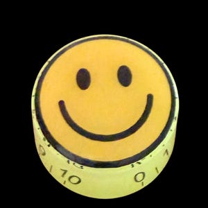 Könnte beinhalten: Gelber Küchenwecker mit einem lächelnden Gesicht. Der Timer hat einen schwarzen Umriss, Augen und Mund. Die Basis des Timers ist mit Zahlen von 0 bis 10 markiert. Der Hintergrund ist schwarz.