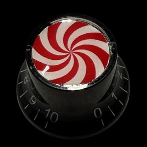 Pode incluir: Botão de guitarra preto com um design em espiral vermelho e branco na parte superior, que lembra uma bala de hortelã. O botão tem números e marcações brancas. O fundo é preto. Acessório para instrumentos musicais.