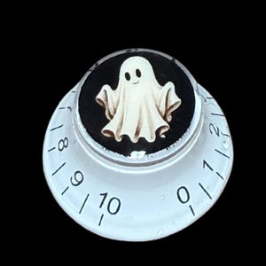 May include: A white circular control knob featuring a black center inlay with a small, cream-colored ghost illustration. The sloped outer edge of the knob is marked with black numbers ranging from zero to ten, indicating a graduated scale for settings.