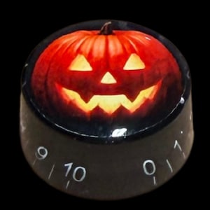 Puede incluir: Un mando de control cilíndrico negro con un gráfico de calabaza de Halloween de color naranja vibrante en la parte superior. El lateral del mando muestra marcas numéricas blancas, incluyendo los números 9, 10 y 0, dispuestos en círculo.
