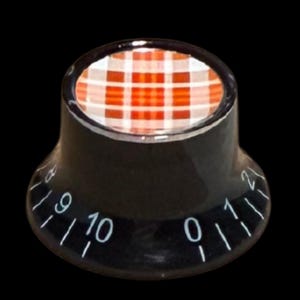 Puede incluir: Un mando de control negro con base acampanada y una inserción circular superior con un patrón de cuadros rojos y blancos. El lateral del mando muestra números impresos en blanco del cero al diez, con marcas entre cada cifra.