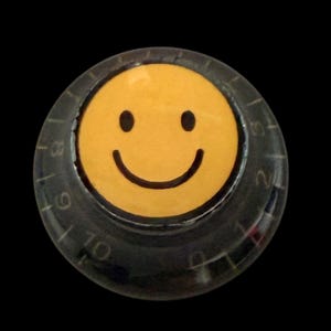 May include: A yellow smiley face button with black eyes and a curved mouth. The button is set within a clear, circular dial with numbers and markings. The background is solid black.