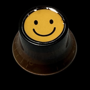 Könnte beinhalten: Ein schwarzer und brauner Bedienknopf mit einem gelben Smiley-Gesicht. Der Knopf hat ein kreisförmiges Design mit schwarzem Rand und brauner Basis. Das Smiley-Gesicht hat schwarze Augen und einen gebogenen Mund.