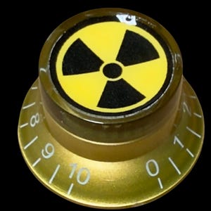 May include: A gold-colored, conical control knob featuring a black and yellow radiation hazard symbol on the top surface. The sloped side of the knob displays white numerical markings ranging from zero to ten, indicating levels of adjustment.