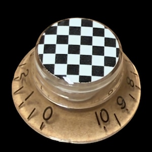 May include: A clear, circular guitar control knob featuring a black and white checkered pattern on the top surface. The outer edge of the knob is transparent and includes black printed numbers ranging from zero to ten for volume or tone adjustment.