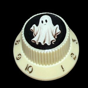 May include: A cream-colored, ribbed control knob featuring a circular top insert with a white ghost illustration on a black background. The side of the knob displays gold-colored numbers ranging from one to ten, arranged in a sequential circular pattern.