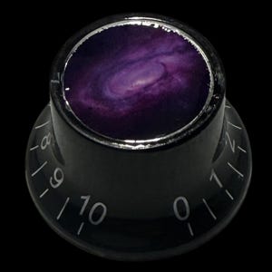 May include: Black guitar control knob with a galaxy design. The knob has a silver rim and white numbers from 0 to 10. The galaxy design features shades of purple and white, resembling a spiral galaxy.