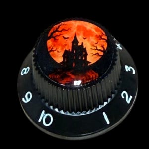 May include: A black circular control knob featuring a top graphic of a dark silhouette of a haunted house against a bright orange full moon. The side of the knob has white printed numbers ranging from one to ten around the outer edge.