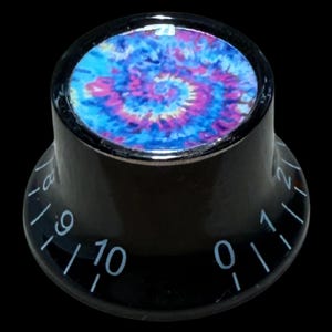 Puede incluir: Un mando de control cilíndrico negro con una inserción circular en la parte superior con un vibrante patrón tie-dye azul, morado y rosa. El lateral del mando está marcado con números blancos impresos del cero al diez para los ajustes.