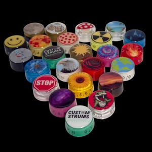 Puede incluir: Una colección de perillas de guitarra con varios diseños, incluyendo una carita sonriente, un hongo, un sol, una rosa y un dinosaurio. Algunas perillas tienen el texto "CUSTOM STRUMS" y números. Las perillas están dispuestas sobre un fondo negro.
