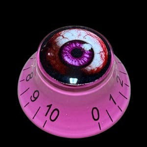 Puede incluir: Un mando de control rosa translúcido con un gráfico circular de un ojo humano realista con un iris morado vibrante y venas rojas. El lado inclinado del mando muestra números negros impresos que van del cero al diez para el ajuste.