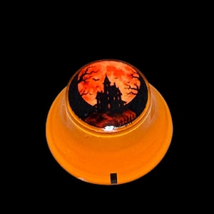 Puede incluir: Un pisapapeles redondo de cristal naranja con la silueta negra de una casa encantada sobre una luna naranja vibrante. El diseño incluye ramas de árboles desnudas y pequeños murciélagos volando, con una base naranja translúcida y una marca negra.