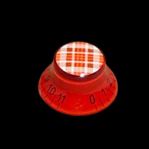 Puede incluir: Un mando de control de guitarra rojo translúcido con una inserción circular en la parte superior con un patrón de cuadros blanco y naranja. El lado inclinado del mando muestra números negros impresos del cero al once para los ajustes.