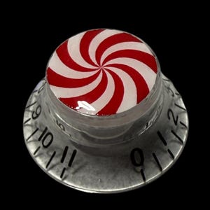 May include: A clear, glass-like knob with a red and white peppermint swirl design on top. The knob has a silver-coloured base with black numbers around the edge, resembling a clock face. The background is black.
