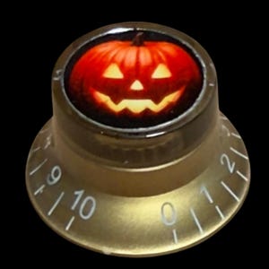 May include: A gold-toned control knob featuring a circular top insert with a glowing orange jack-o'-lantern graphic. The sloped side of the knob displays white printed numbers ranging from zero to ten, indicating a standard volume or tone dial.