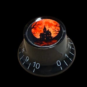 May include: A black cylindrical control knob featuring a circular top insert with a graphic of a dark silhouette of a haunted house against a bright orange full moon. The side of the knob displays white printed numbers ranging from zero to ten.