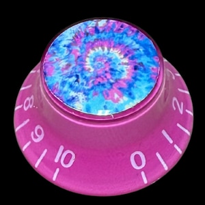 Puede incluir: Un mando de control rosa con una inserción circular en la parte superior con un patrón tie-dye azul y morado. El lado inclinado del mando está marcado con números blancos del cero al diez, indicando una escala graduada para ajuste o volumen.
