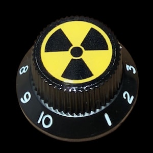 May include: A black circular control knob with a ribbed edge and a flat top featuring a yellow and black radiation warning symbol. The base of the knob is marked with white printed numbers ranging from one to ten in a clockwise sequence.