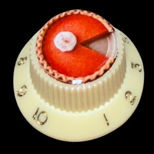 May include: A cream-colored cylindrical control knob featuring a fluted side and gold-colored numbers from one to ten printed around the base. The top surface displays a graphic of an orange pumpkin pie with a single white dollop of whipped cream.