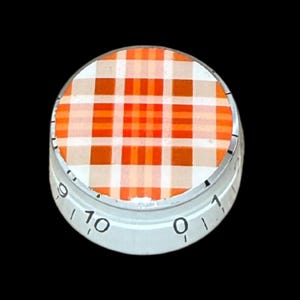Op de afbeelding: Een ronde bedieningsknop met een levendig oranje, wit en beige ruitpatroon op het bovenoppervlak. De zijkant van de witte knop is gemarkeerd met zwarte cijfers van nul tot tien, wat een schaalverdeling aangeeft voor een nauwkeurige afstelling.