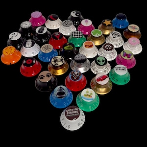 Puede incluir: Una colección de perillas de guitarra en varios colores y diseños, incluyendo un símbolo de yin-yang, una huella de pata y un coche rojo. Algunas perillas tienen el texto "CUSTOM STRUMS" y números.