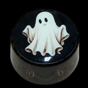 Puede incluir: Un mando de control de guitarra cilíndrico negro con una ilustración de un fantasma blanco en la superficie superior. El lateral del mando muestra marcas numéricas que van del cero al diez en una fuente dorada, ofreciendo una escala visual.