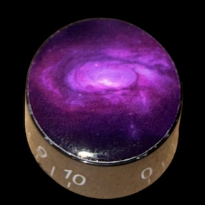 Könnte beinhalten: Runder Knopf mit einem lila und rosa Galaxiemuster. Der Knopf hat einen silbermetallischen Rand und eine beige Basis mit weißen Zahlen und Markierungen. Dieser Knopf ist wahrscheinlich für eine Gitarre oder ein anderes elektronisches Gerät.