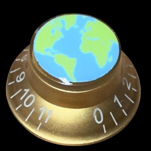 Pode incluir: Um botão de controlo circular de cor dourada com uma inserção plana no topo que apresenta um mapa do mundo em azul e verde claro. O lado inclinado do botão exibe números brancos impressos de zero a onze com marcas de graduação.