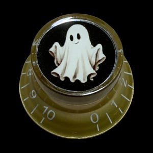 Puede incluir: Un mando de control de guitarra circular dorado con una incrustación central negra que presenta una ilustración de un fantasma blanco. El mando tiene un acabado translúcido, un borde superior plateado y marcas numéricas blancas de cero a diez.