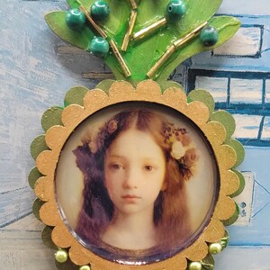 May include: A decorative wall hanging featuring a portrait of a young woman with long hair and flowers. The portrait is framed with a scalloped gold and green border, adorned with green beads. Above the portrait is a green leaf design with green beads.