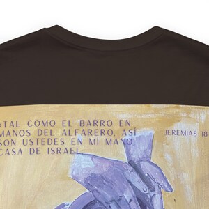 以下が含まれることがあります： 黒いTシャツに金と白で描かれたデザイン。デザインにはスペイン語のテキストと手の描写が含まれています。テキストは「TAL COMO EL BARRO EN MANOS DEL ALFARERO, ASÍ SON USTEDES EN MI MANO, CASA DE ISRAEL」と「JEREMIAS 18:」と書かれています。